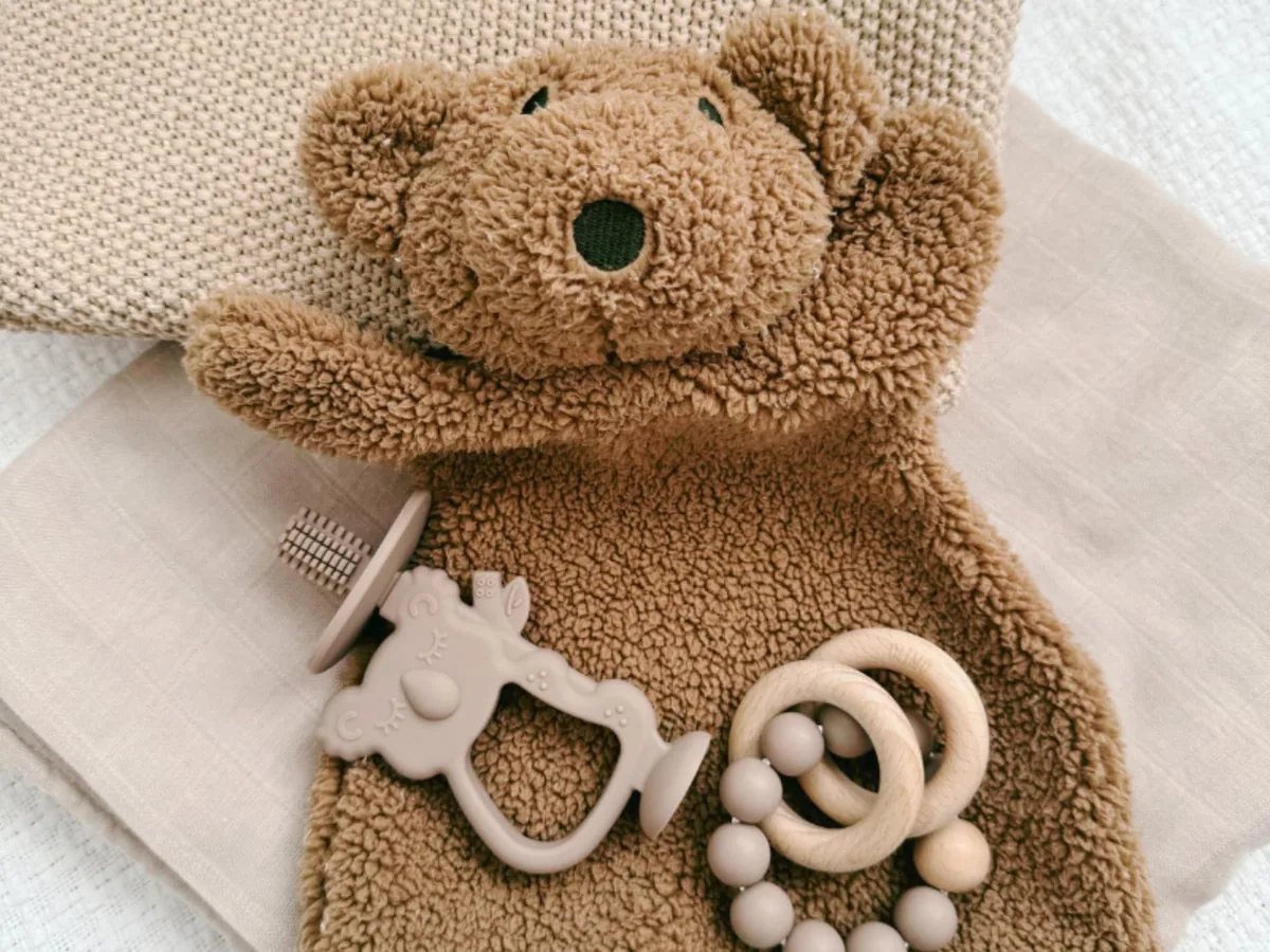 A teddy bear comforter laid out with a silicone teether and a wooden rattle ring